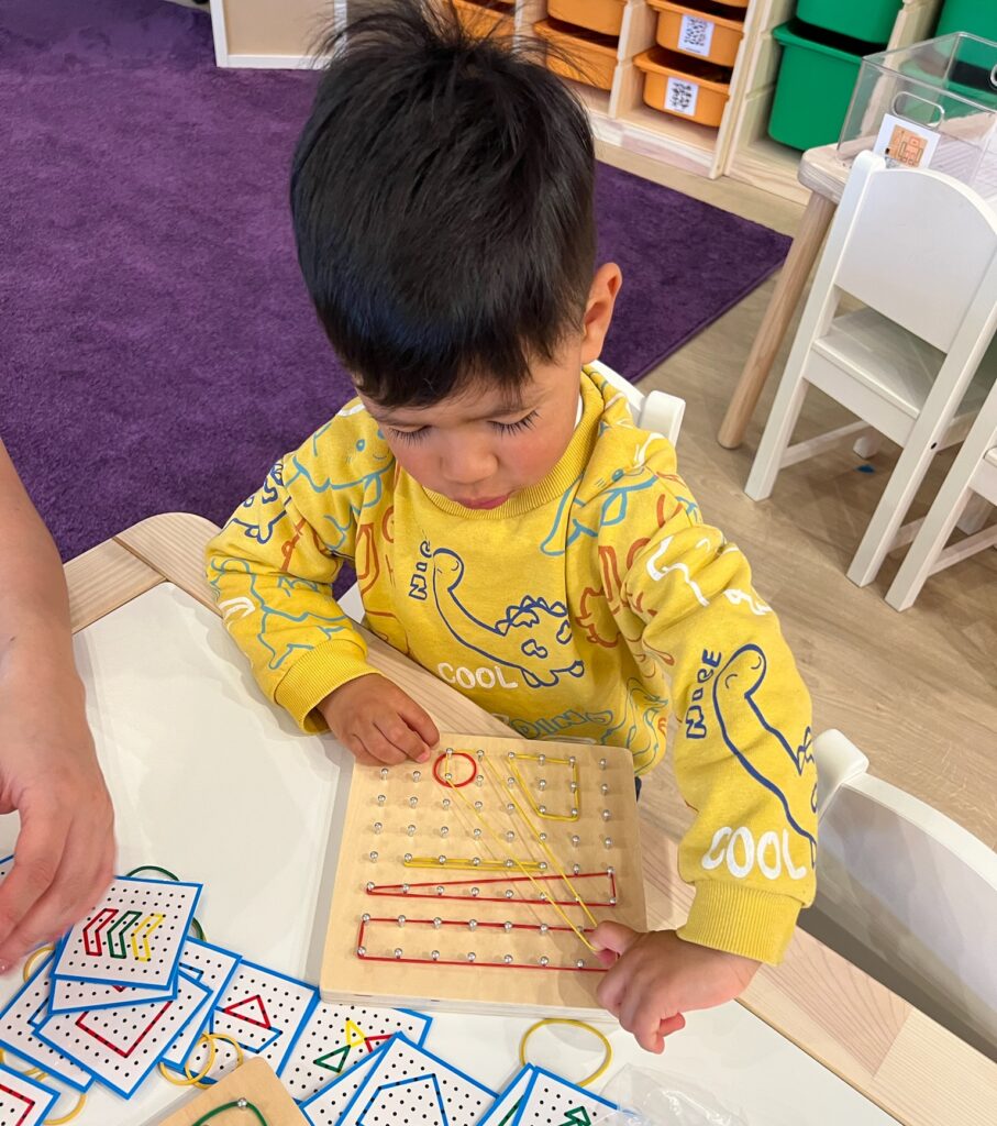 A young boy in a yellow sweatshirt carefully stretches colored rubber bands across a wooden geoboard to match geometric pattern cards on the table.
