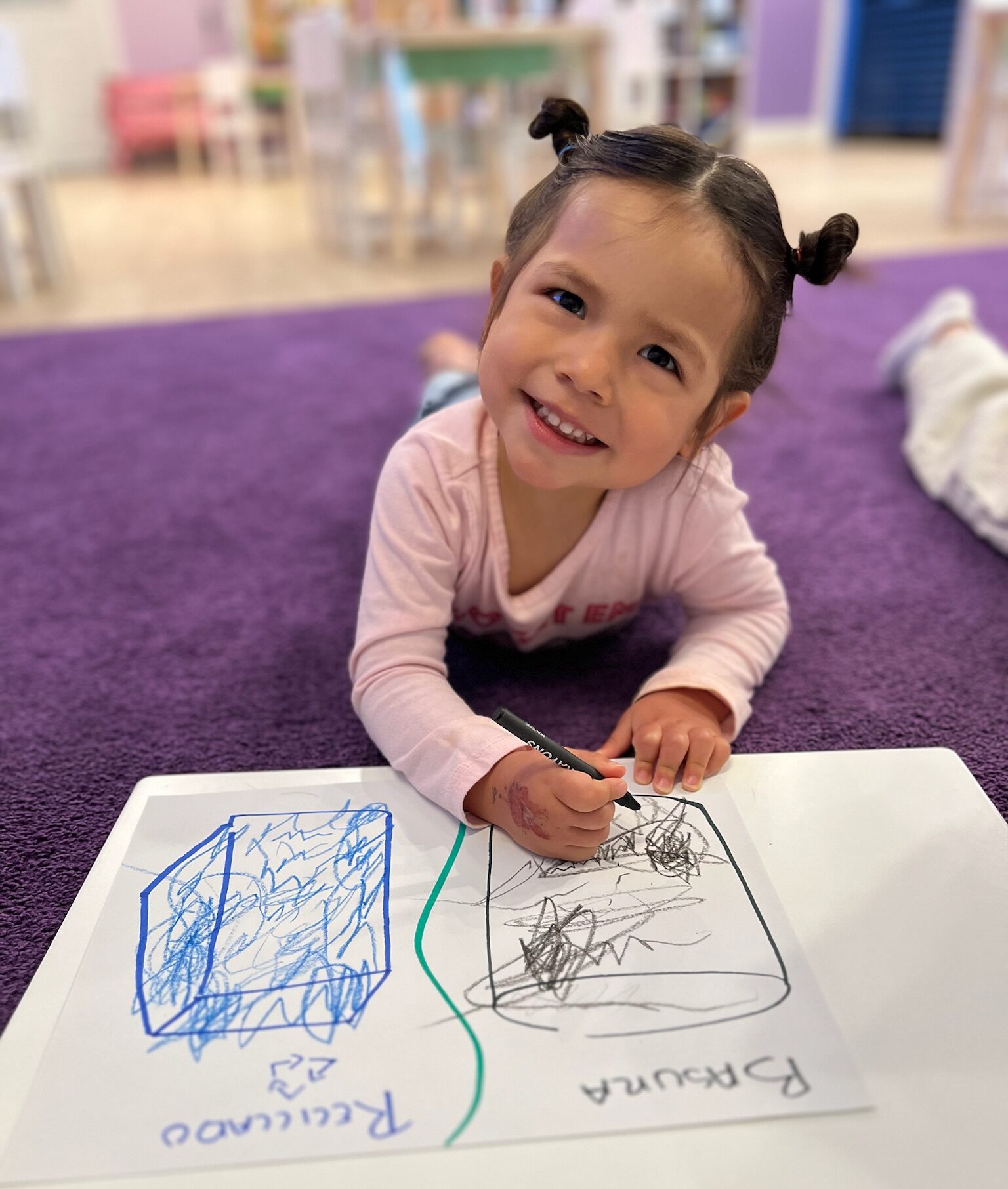 A smiling young preschool girl lying on a purple rug, coloring a Spanish recycling and trash activity sheet with markers